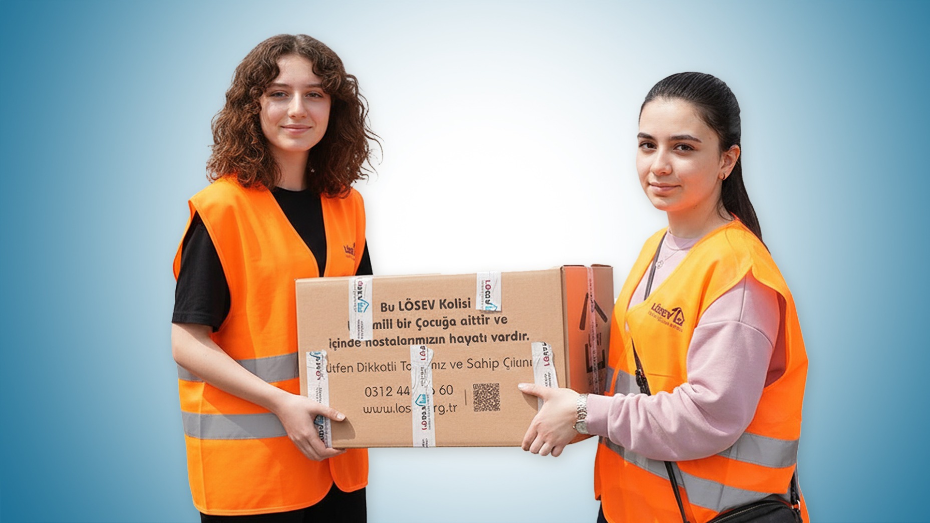 İki LÖSEV gönüllüsü kadın, ellerinde üzerinde “Bu LÖSEV Kolisi lösemili bir çocuğa aittir” yazan yardım kolisini taşıyor.